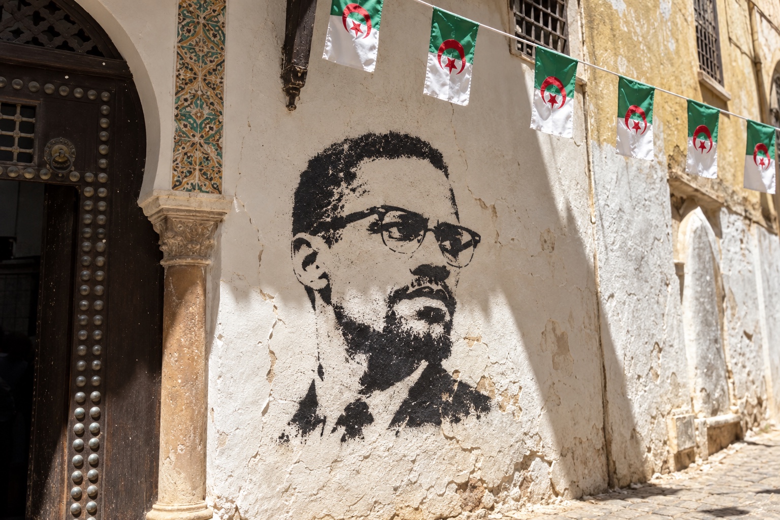 Composition artistique : portrait de Malcolm X au pochoir sur un mur de la Casbah, sous des drapeaux algériens.